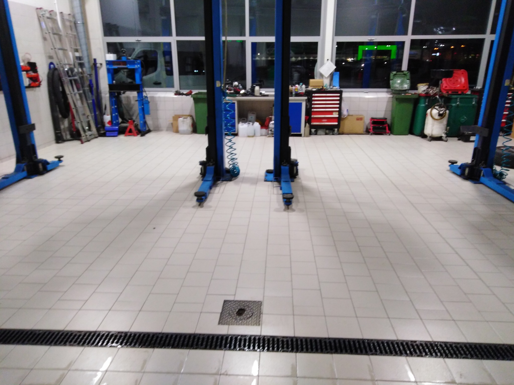 Czysta, wykafelkowana posadzka w warsztacie samochodowym z czterema podnośnikami kolumnowymi, widoczne narzędzia, drabiny i pojemniki na śmieci w tle.