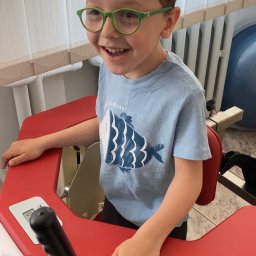 Fizjo Kraina. Rehabilitacja dzieci i niemowląt Monika Kalińska - Uśmiechnięty chłopiec w okularach siedzi na czerwonym urządzeniu rehabilitacyjnym z uchwytami, na tle białych zasłon i grzejnika