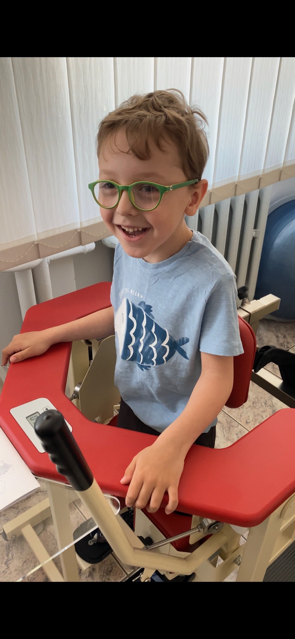 Uśmiechnięty chłopiec w okularach siedzi na czerwonym urządzeniu rehabilitacyjnym z uchwytami, na tle białych zasłon i grzejnika