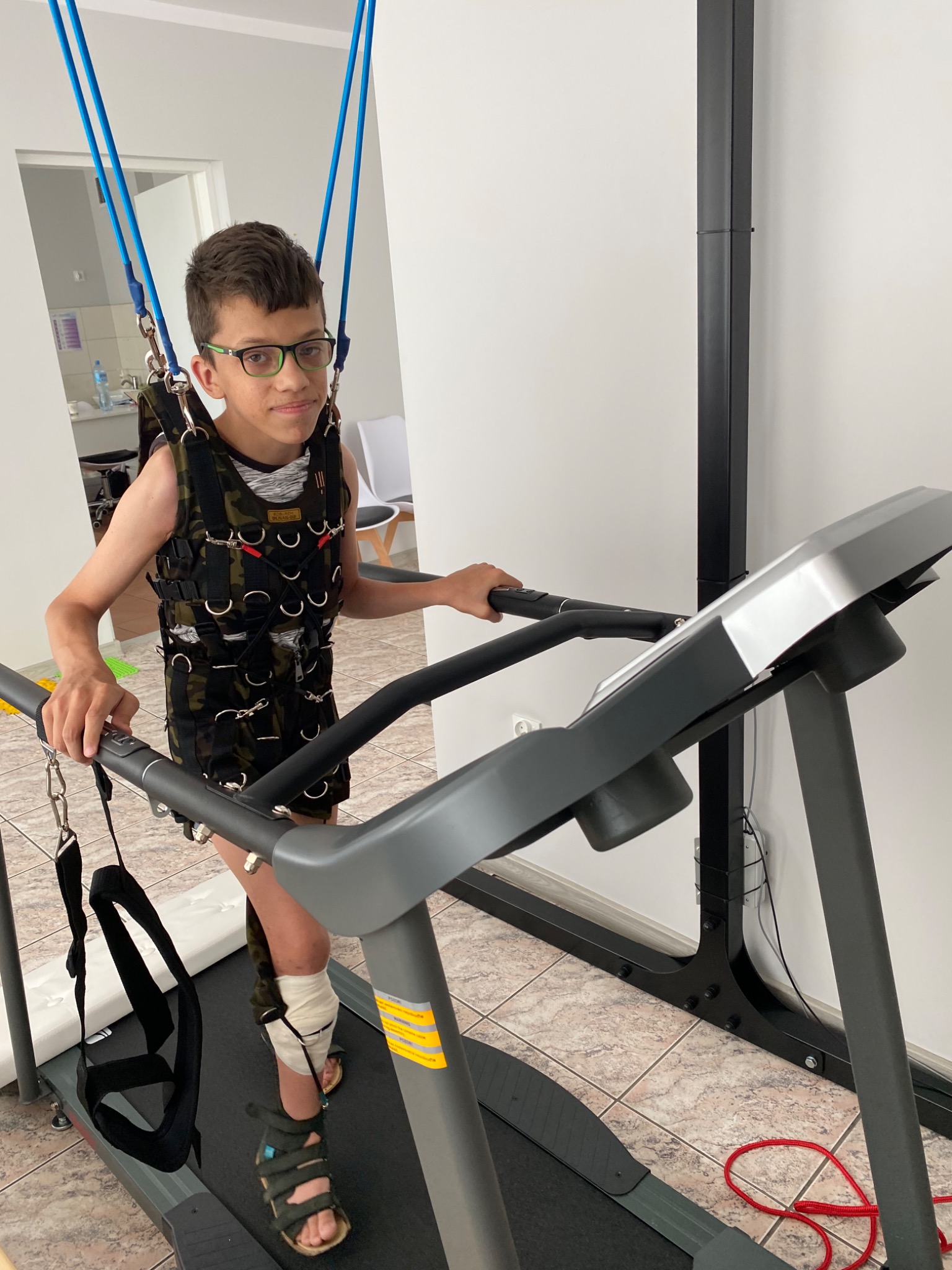 Chłopiec w okularach, w kamizelce i z zabandażowaną nogą, ćwiczy na bieżni rehabilitacyjnej podwieszony na linach.