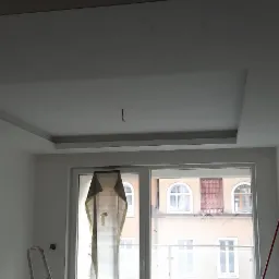 Wykończone wnętrze z podwieszanym sufitem w kształcie kwadratu z oświetleniem, widok na okno z zasłoniętą częścią szyby, drabina i czerwony kij oparty o ścianę.