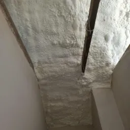 Poddasze ocieplone białą pianką poliuretanową, widoczne drewniane elementy konstrukcyjne i fragment ściany.