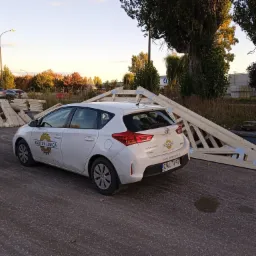 Biały samochód osobowy z logo firmy 'Tartak Izbica' na tle drewnianych konstrukcji dachowych i stosu tarcicy na placu.