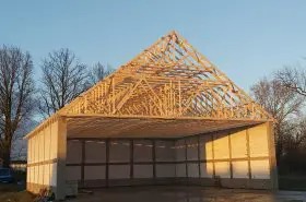 Konstrukcja szkieletowa domu w trakcie budowy, widoczne drewniane elementy więźby dachowej i ściany z izolacją, w tle błękitne niebo o zachodzie słońca.