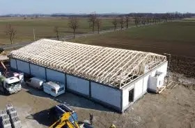 Wznoszenie konstrukcji budynku z prefabrykatów, widoczna drewniana więźba dachowa, w tle krajobraz polny i droga.