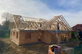 Wznoszenie drewnianej konstrukcji dachu na murowanym domu jednorodzinnym w trakcie budowy, widoczne elementy więźby dachowej, praca na wysokości, częściowo otynkowane ściany, krajobraz wiejski w tle.