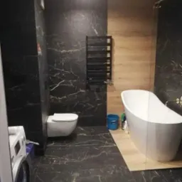 Nowoczesna łazienka z czarnymi płytkami imitującymi marmur, wolnostojącą białą wanną i drewnianymi panelami ściennymi w strefie prysznicowej.