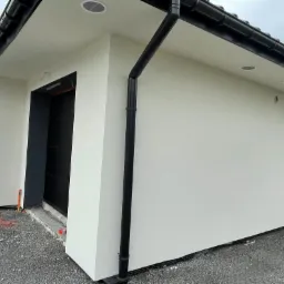 Nowoczesny dom jednorodzinny z białą elewacją o strukturze baranka, czarną stolarką okienną i dachem z ciemnej dachówki, widok z poziomu gruntu.