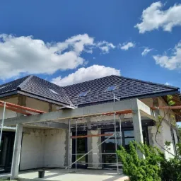 Dom w stanie surowym otwartym, widoczne białe bloczki, betonowe słupy, czarna dachówka z oknami dachowymi i rusztowania, błękitne niebo z chmurami w tle.