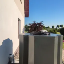 Zewnętrzna jednostka pompy ciepła ustawiona na betonowych płytach obok ściany domu, z rurami przyłączeniowymi i ozdobnym drzewem na tle błękitnego nieba.