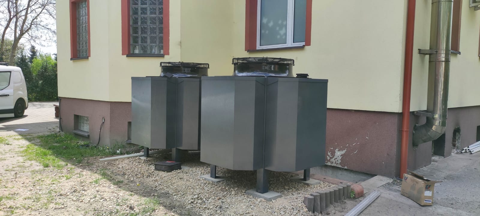 Dwie szare jednostki zewnętrzne z wentylatorami na górze, stojące na kamienistym podłożu przy żółtej ścianie budynku z widocznymi oknami i rurą spustową.