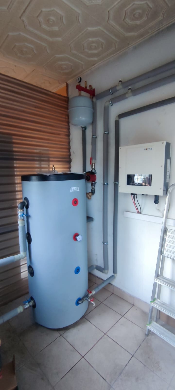 Szary zbiornik buforowy z podłączonymi rurami i zaworami, obok falownik solarny na ścianie, drabina w tle, w pomieszczeniu z kafelkami na podłodze i dekoracyjnym sufitem.