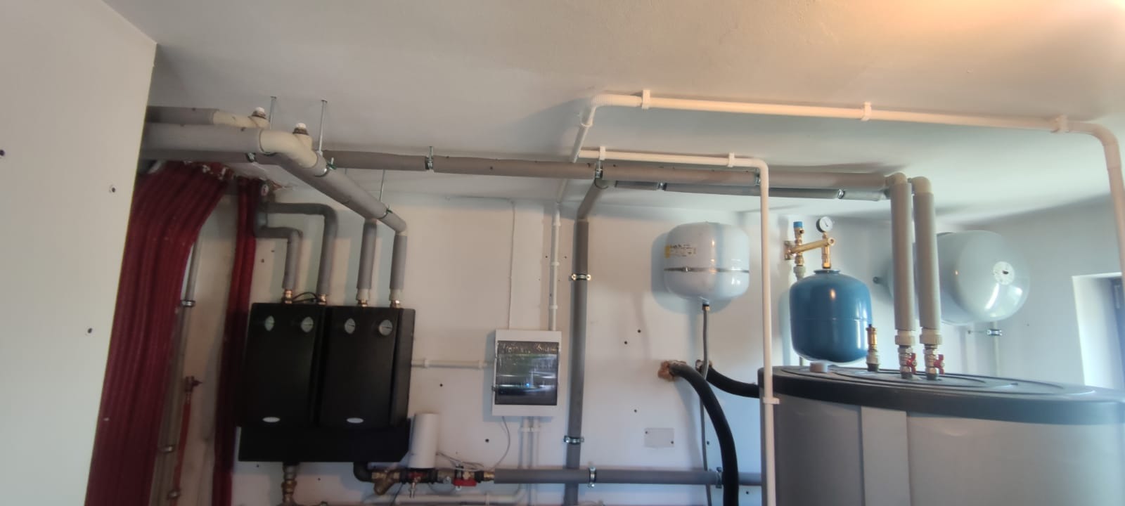 Kompletna instalacja grzewcza w kotłowni z dwoma czarnymi modułami grzewczymi, zbiornikami wyrównawczymi, rozbudowanym systemem rur i przewodów, oraz skrzynką elektryczną na ścianie.