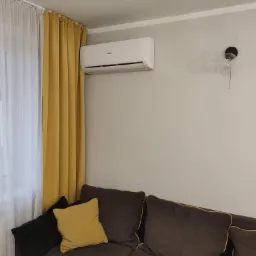 Biały klimatyzator Haier zamontowany na ścianie w salonie, nad brązową sofą z żółtymi poduszkami, obok okna z białymi i żółtymi zasłonami oraz lampy ściennej z żarówką na sznurku.