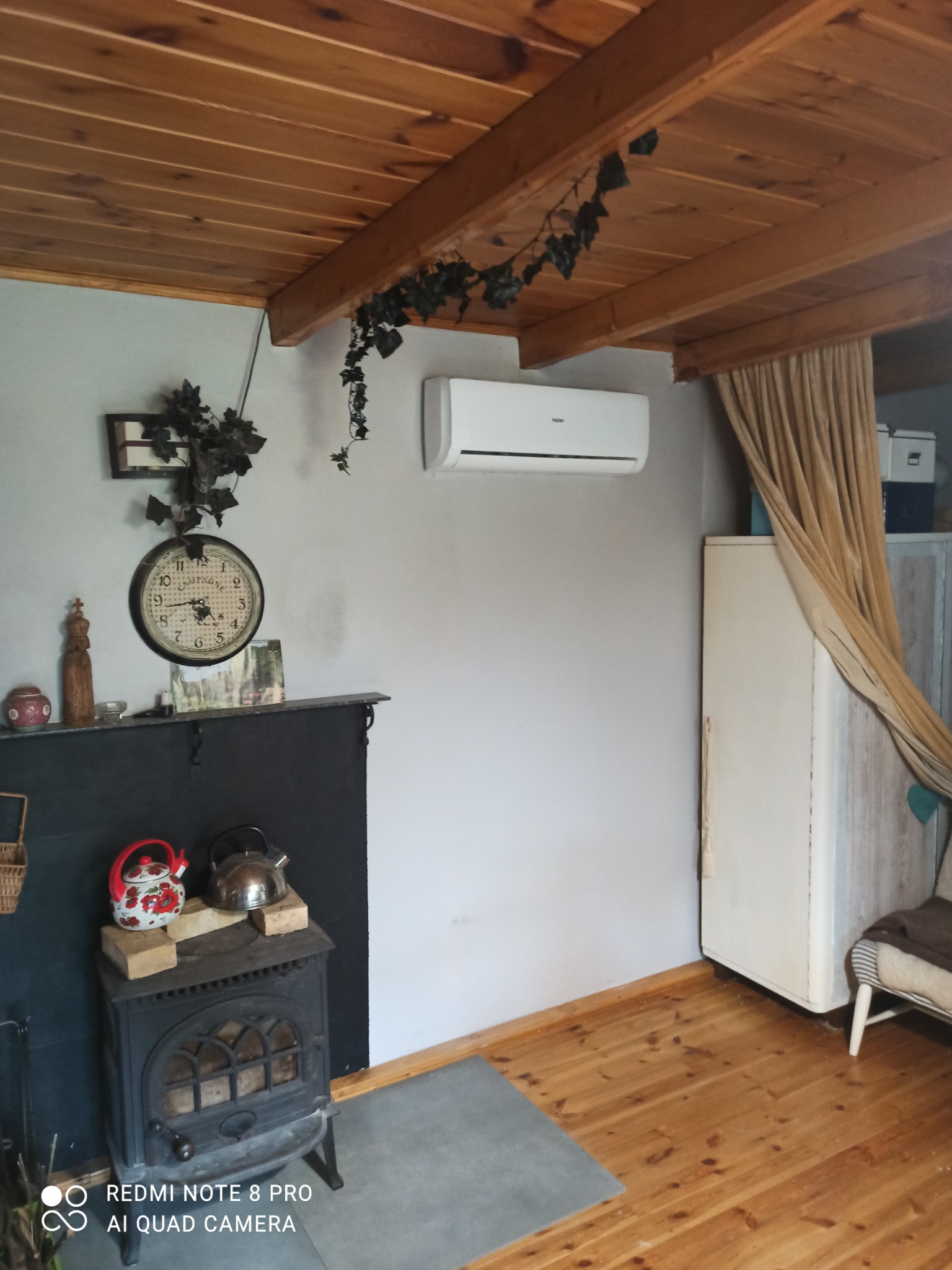 Wnętrze pokoju z drewnianym sufitem, na ścianie zamontowany klimatyzator marki Haier, poniżej dekoracyjny kominek z czajnikami, zegar i ozdobne pnącza.