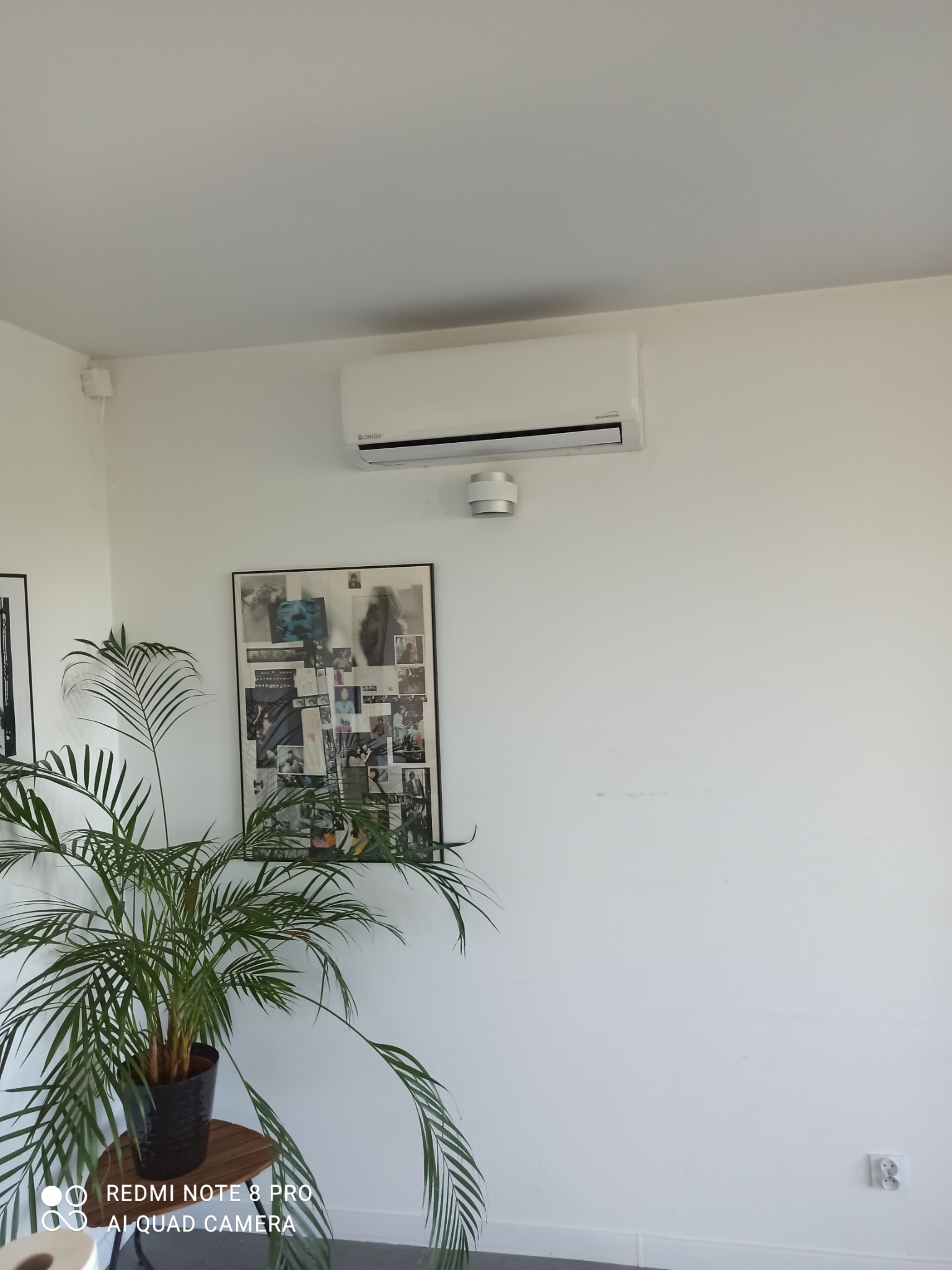 Biały klimatyzator ścienny marki CHIGO zamontowany w jasnym pomieszczeniu, widoczny czujnik dymu, plakat i roślina doniczkowa.