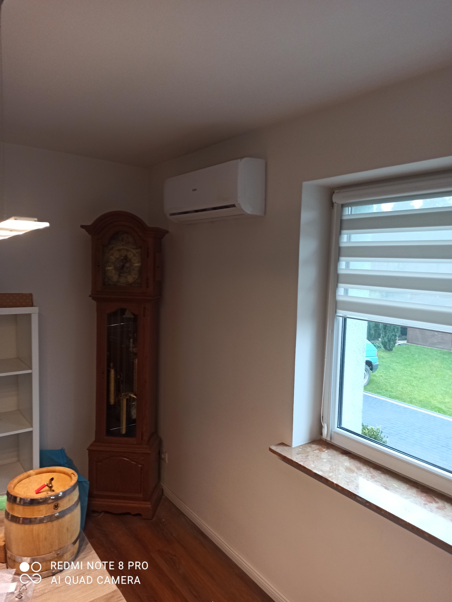 Wnętrze pokoju z zainstalowanym klimatyzatorem ściennym, widok na okno z roletami dzień-noc i zabytkowy zegar stojący.