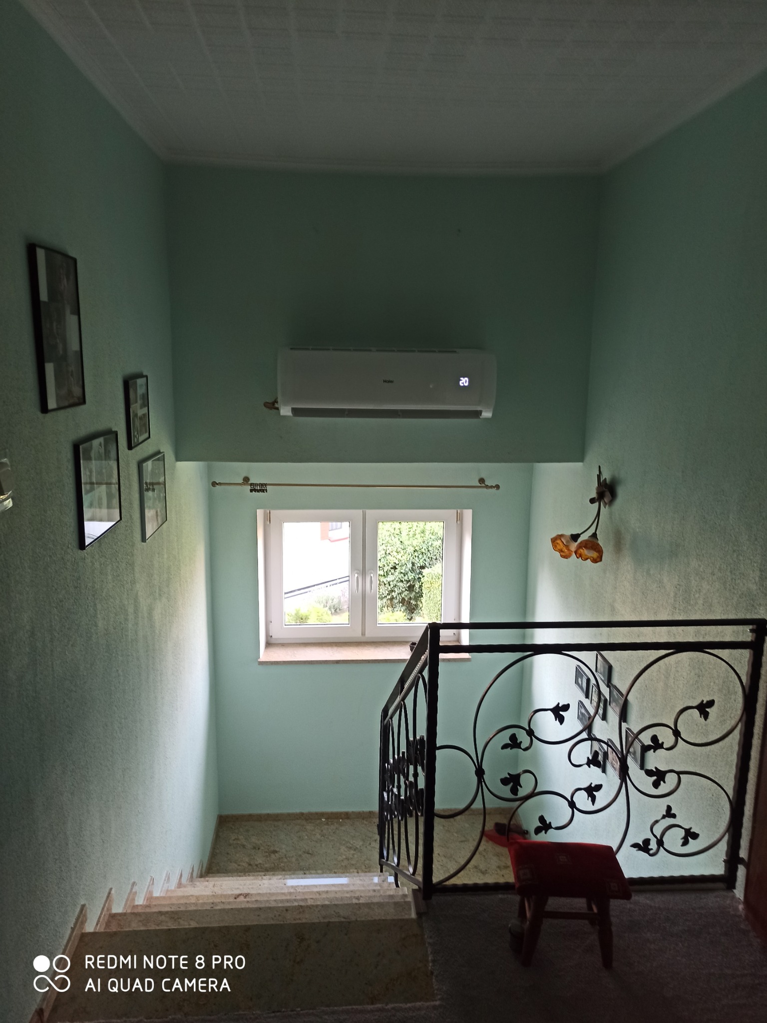 Korytarz z klimatyzatorem Haier zamontowanym nad oknem, widok ze schodów w dół, ściany w odcieniach zieleni, dekoracyjna balustrada z motywem roślinnym.