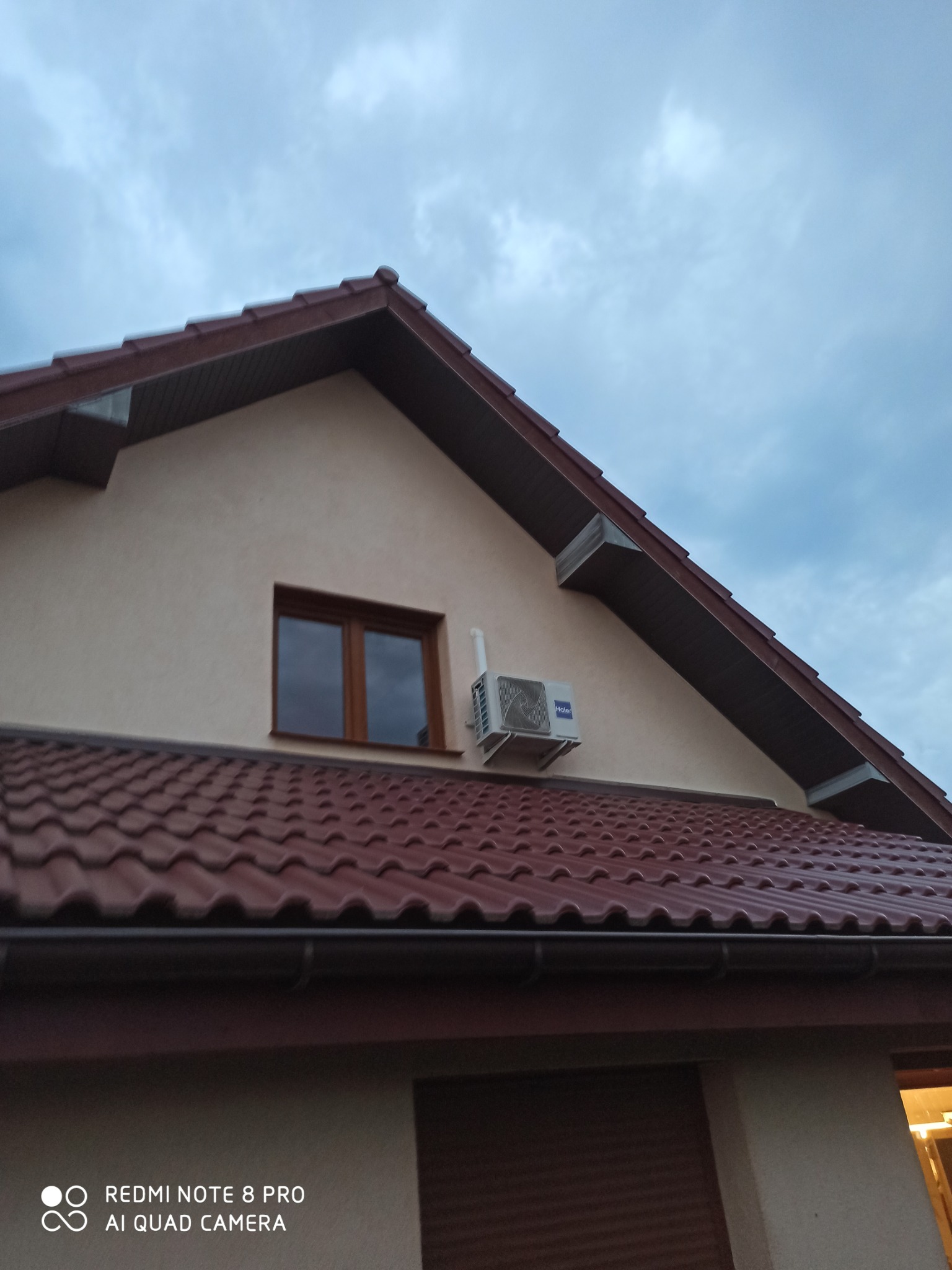 Zewnętrzna jednostka klimatyzatora marki Haier zamontowana na ścianie szczytowej domu jednorodzinnego, widok z dołu na tle pochmurnego nieba.