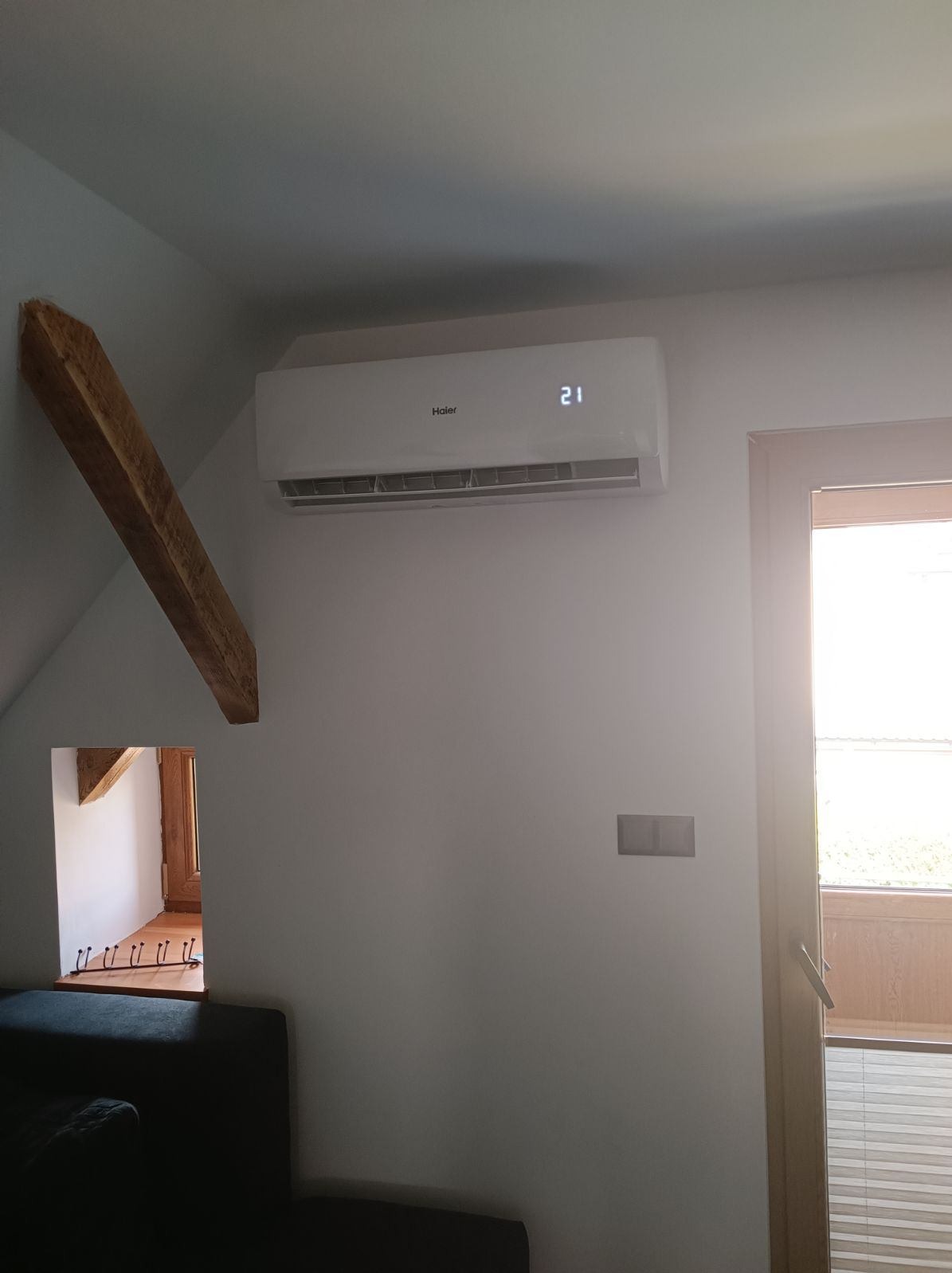 Biały klimatyzator Haier zamontowany na ścianie w pokoju na poddaszu, obok widoczna drewniana belka stropowa i okno balkonowe.