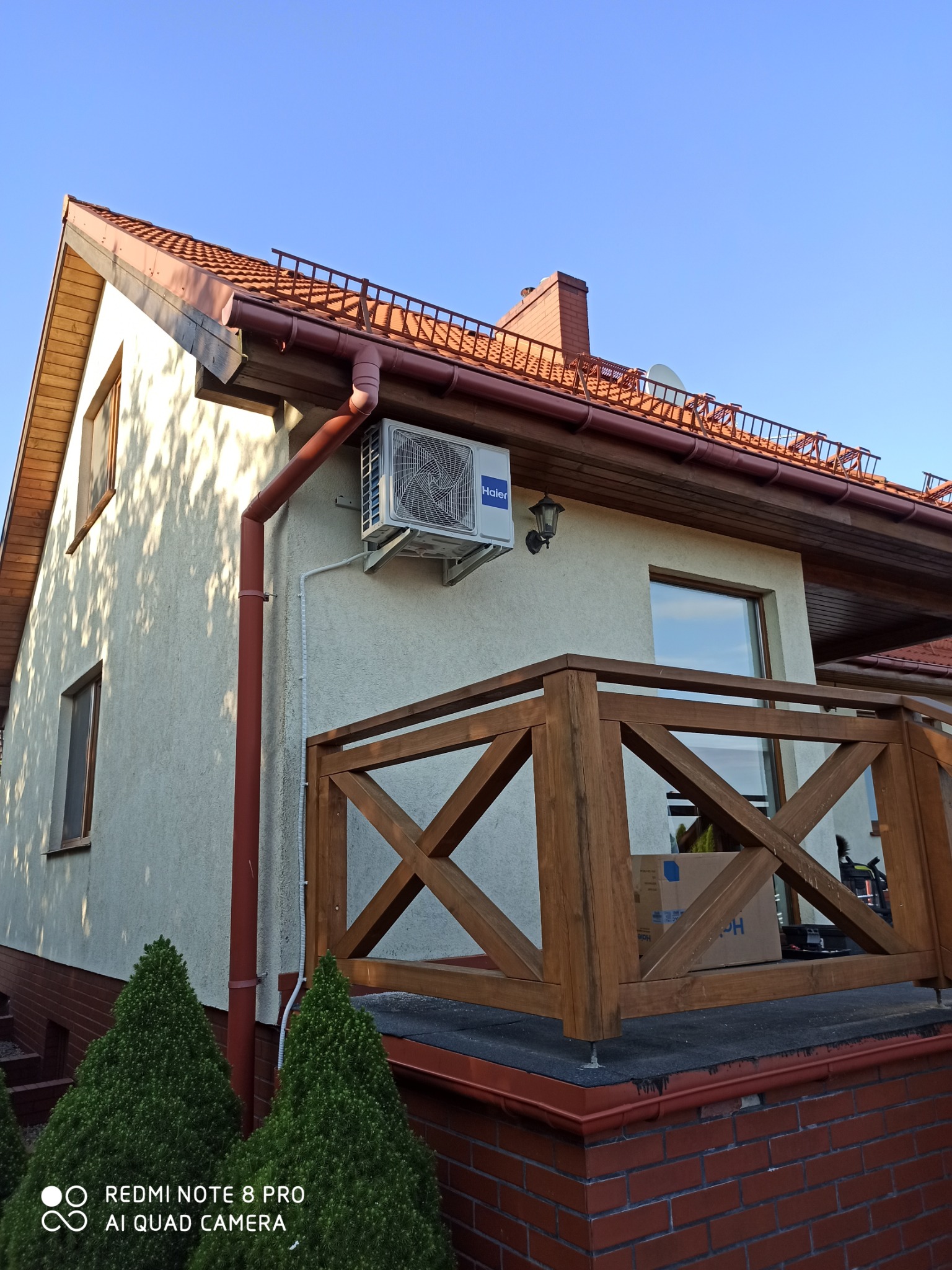Zewnętrzna jednostka klimatyzacji Haier zamontowana na elewacji domu z drewnianą balustradą balkonu, widok z dołu w słoneczny dzień.