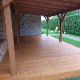 Drewniana altana z brązowym zadaszeniem i podłogą, ściana wykończona kamieniem dekoracyjnym, w tle zielony żywopłot i leżak.