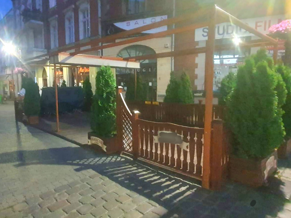 Zewnętrzna drewniana konstrukcja z zadaszeniem i ozdobnym płotkiem, otaczająca przestrzeń przed budynkiem z szyldem 'Salon' i 'Studio Filmowe', udekorowana tujami w donicach, oświetlona wieczornym...