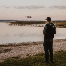 Mężczyzna w czapce obserwuje drona unoszącego się nad jeziorem z drewnianym pomostem i trzcinami w tle, podczas zachodu słońca.