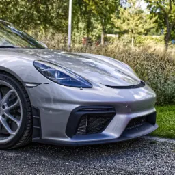 Srebrny sportowy samochód Porsche 718 Cayman GT4 RS z czerwonym dachem na tle zieleni, widok z przodu, z delikatnym logo 'Drone Effect' w lewym dolnym rogu.