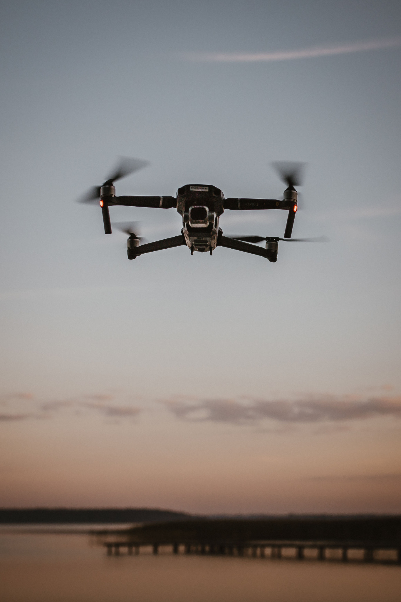 Czarny dron typu Mavic 2 w locie na tle zachodzącego słońca nad wodą i pomostem, z widocznymi czerwonymi światłami pozycyjnymi.