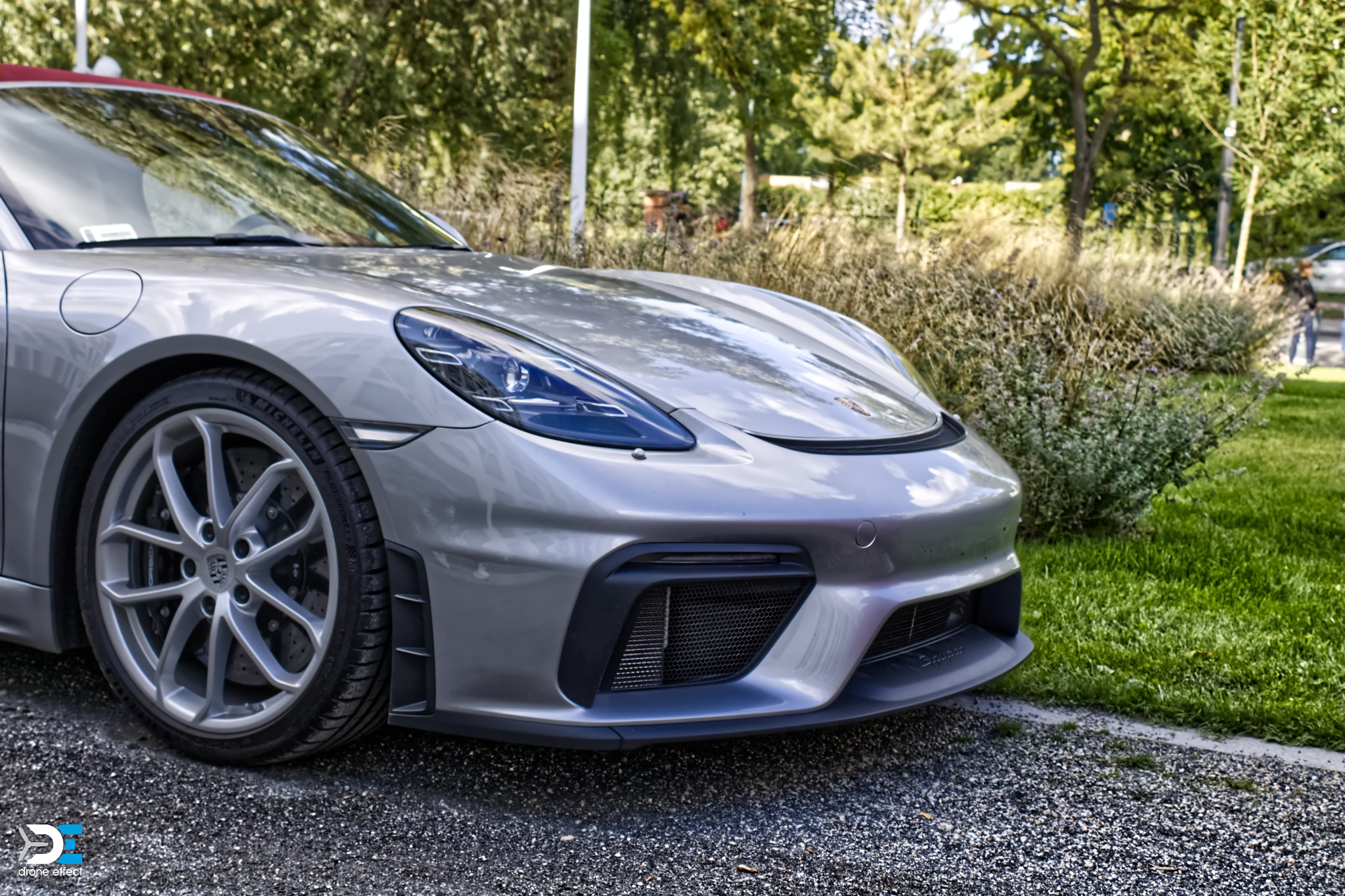 Srebrny sportowy samochód Porsche 718 Cayman GT4 RS z czerwonym dachem na tle zieleni, widok z przodu, z delikatnym logo 'Drone Effect' w lewym dolnym rogu.