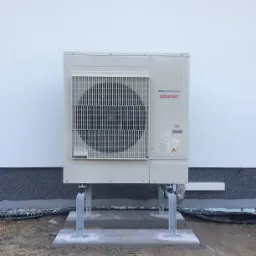 Zewnętrzna jednostka pompy ciepła Atlantic DC Inverter zamontowana na betonowym fundamencie i metalowych wspornikach przy białej ścianie budynku z szarą podmurówką.