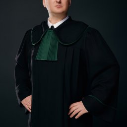 KANCELARIA ADWOKACKA SIEŃKO PIĘTA I PARTNERZY SPÓŁKA CYWILNA - Portret mężczyzny w todze adwokackiej, z rękami opartymi na biodrach, na ciemnym tle.