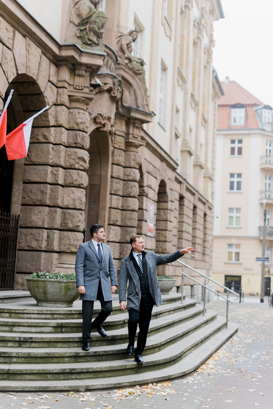 Dwóch mężczyzn w płaszczach schodzi po schodach kamiennego budynku z flagą Polski, jeden wskazuje palcem