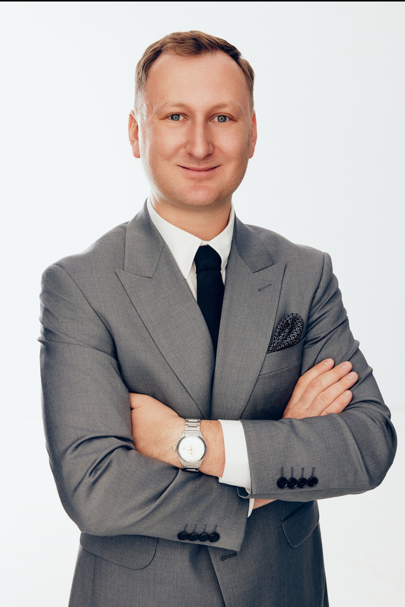 Portret adwokata z Poznania w szarym garniturze, z rękami skrzyżowanymi na piersiach i srebrnym zegarkiem na nadgarstku, na białym tle.