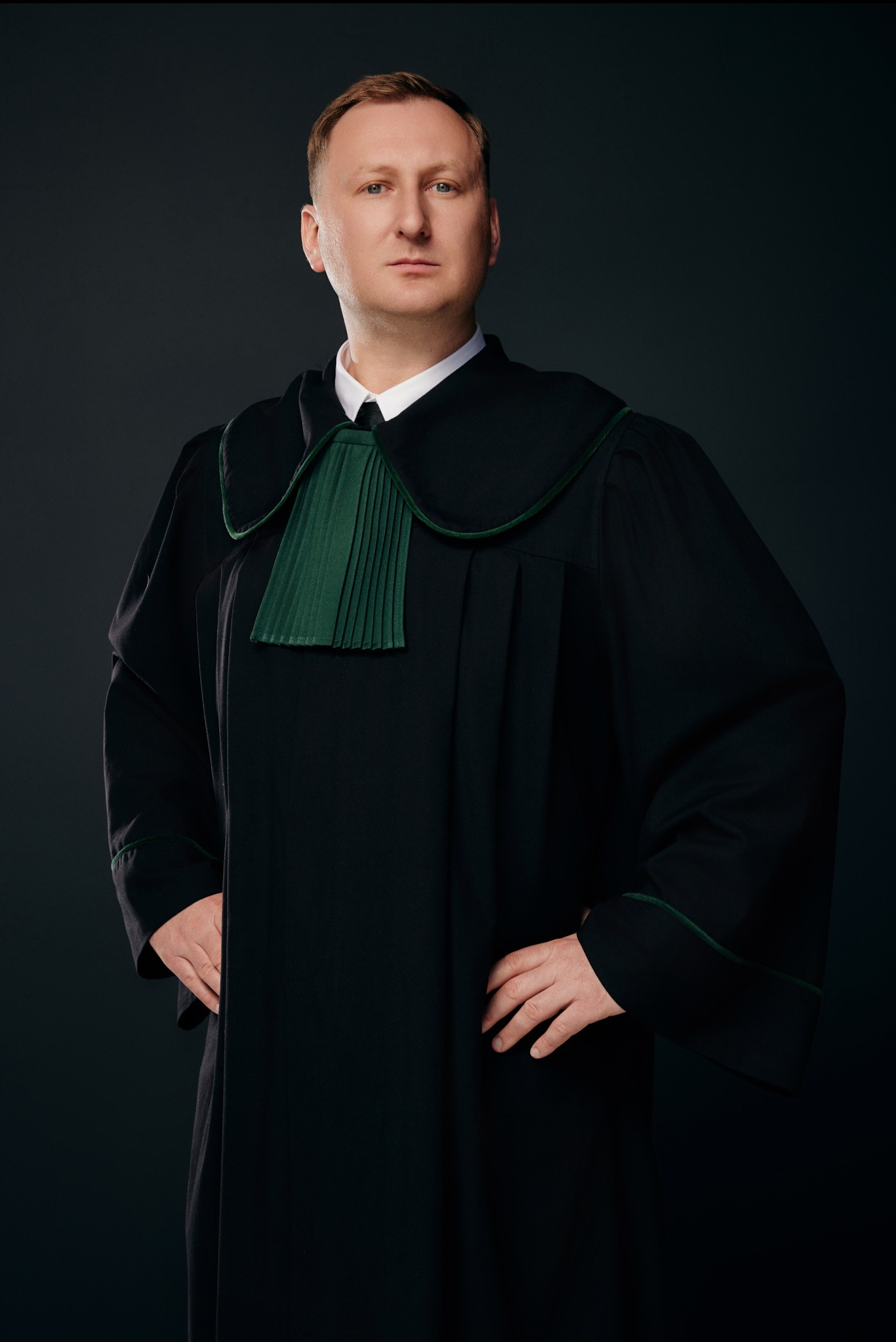 Portret mężczyzny w todze adwokackiej, z rękami opartymi na biodrach, na ciemnym tle.