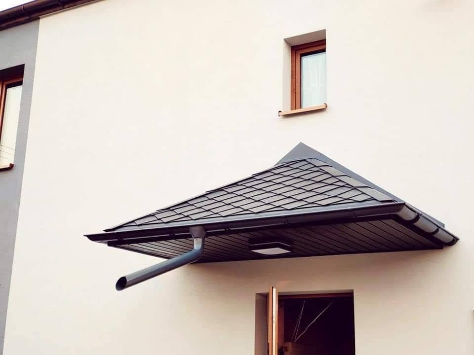Nowoczesne zadaszenie wejścia z grafitową dachówką bitumiczną, rynną spustową i oświetleniem punktowym, zamontowane nad drzwiami wejściowymi do budynku o jasnej elewacji.