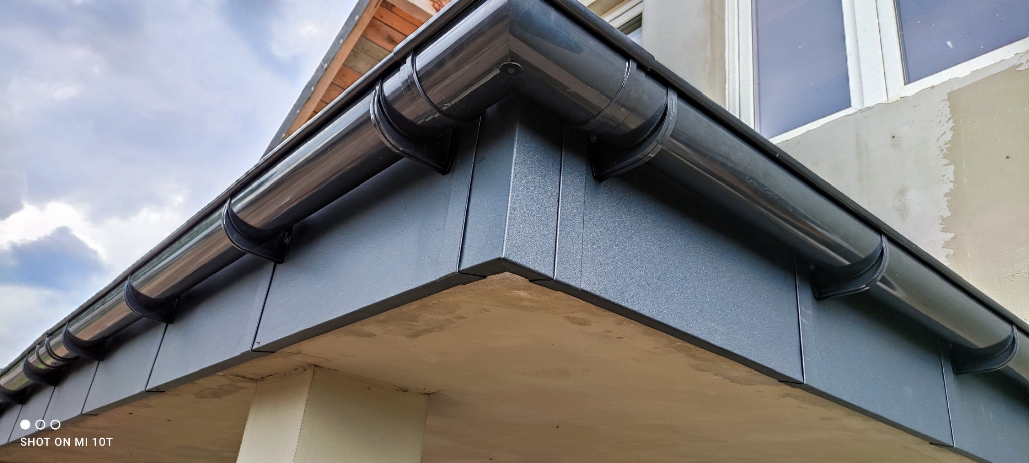Nowo zamontowane, błyszczące, grafitowe rynny z widocznymi uchwytami, zamocowane do ciemnoszarej podbitki dachowej na tle fragmentu elewacji budynku w trakcie wykończenia.