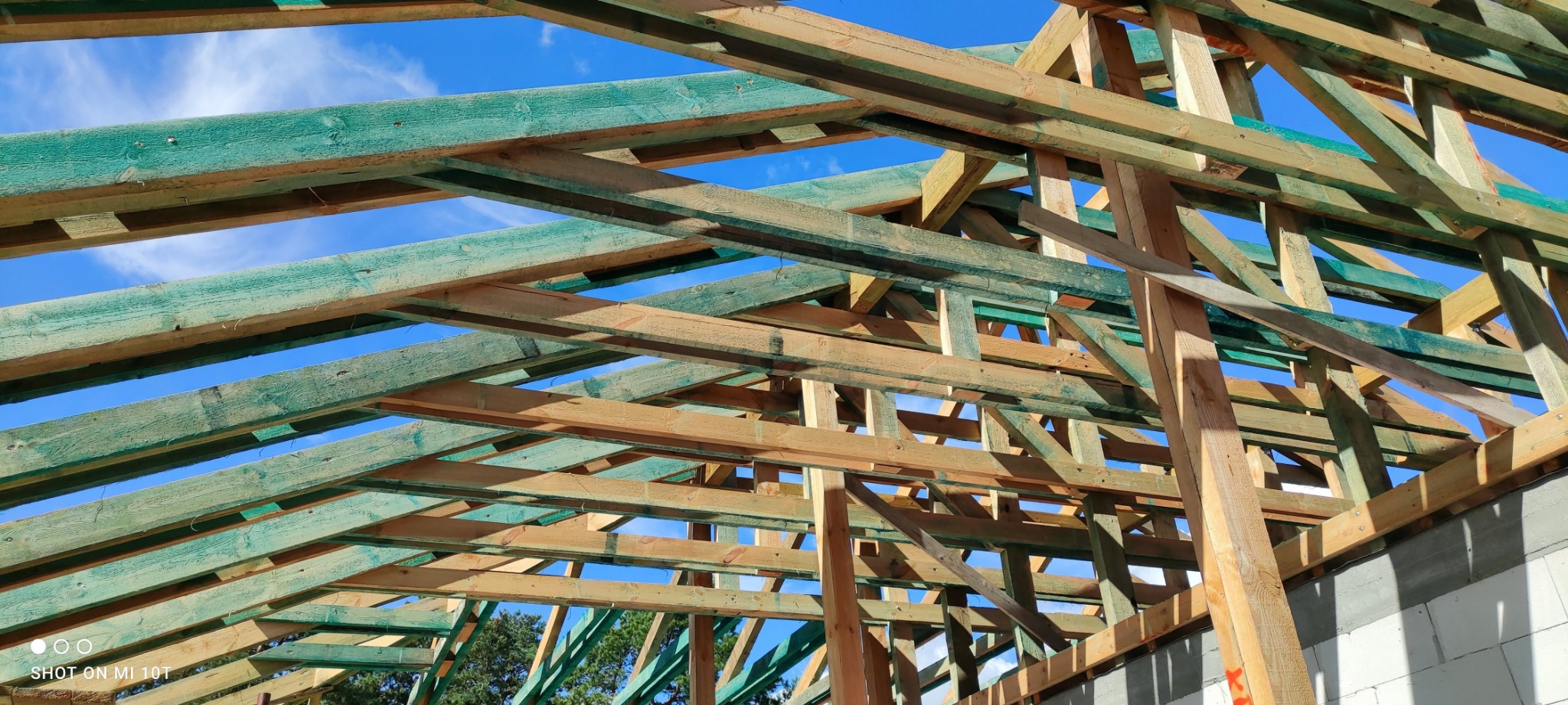Konstrukcja drewniana dachu w trakcie budowy, impregnowane elementy w kolorze zielonym, widok od dołu na tle błękitnego nieba.