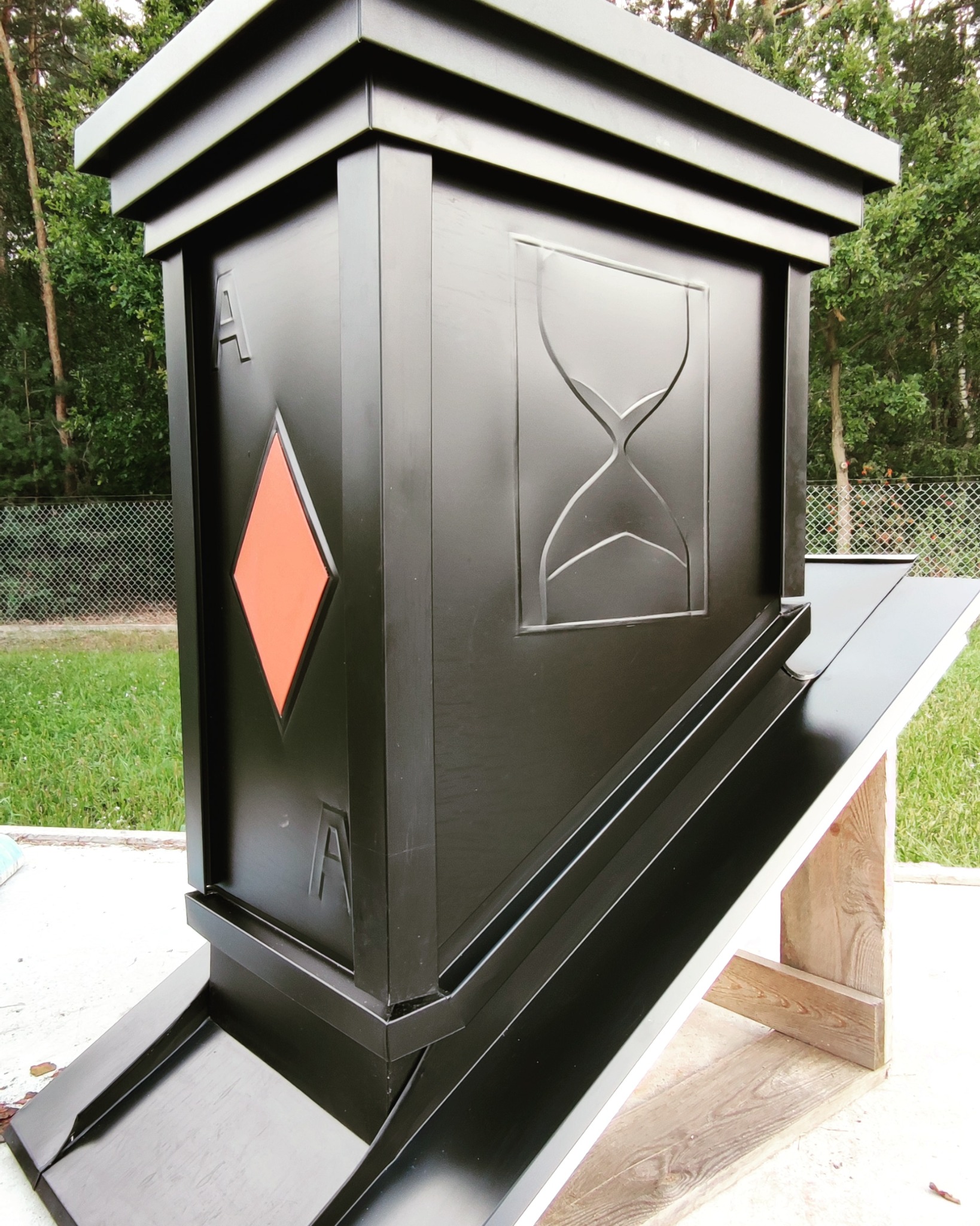 Czarna, metalowa obudowa rynny z motywem karo i klepsydry, zamontowana na drewnianej konstrukcji. Widoczne elementy systemu odprowadzania wody.