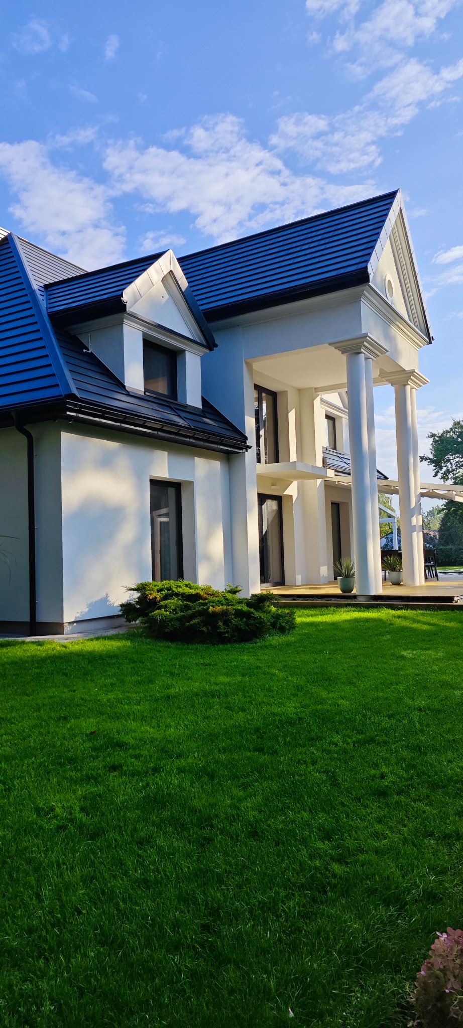 Elegancki, dwukondygnacyjny dom z ciemnym dachem i białymi kolumnami, otoczony zielonym trawnikiem w słoneczny dzień. Widoczne okna i elementy architektoniczne.