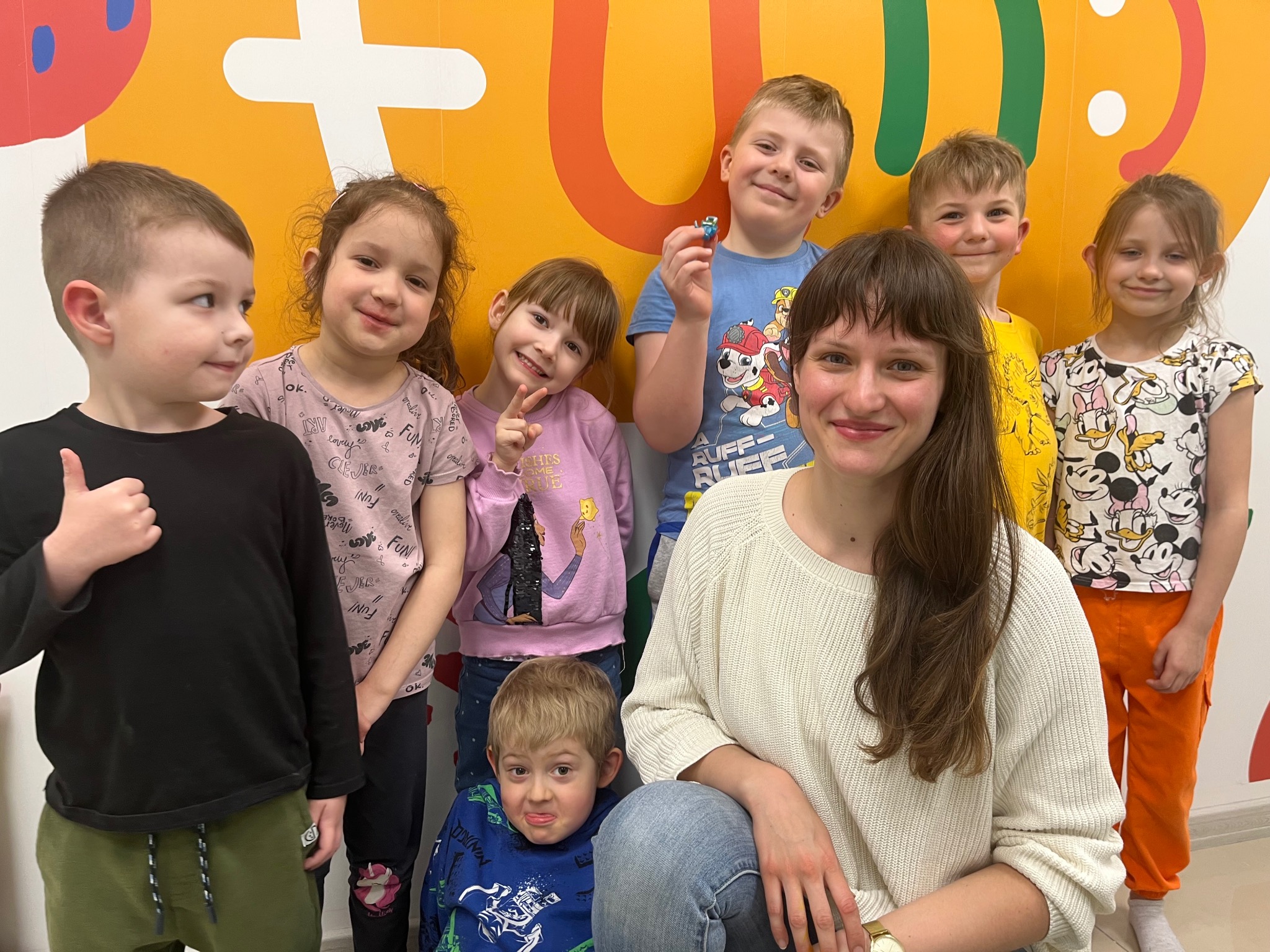 Uśmiechnięta nauczycielka z grupą dzieci na tle kolorowej ściany z motywami edukacyjnymi, tworząca przyjazną atmosferę w klasie językowej.