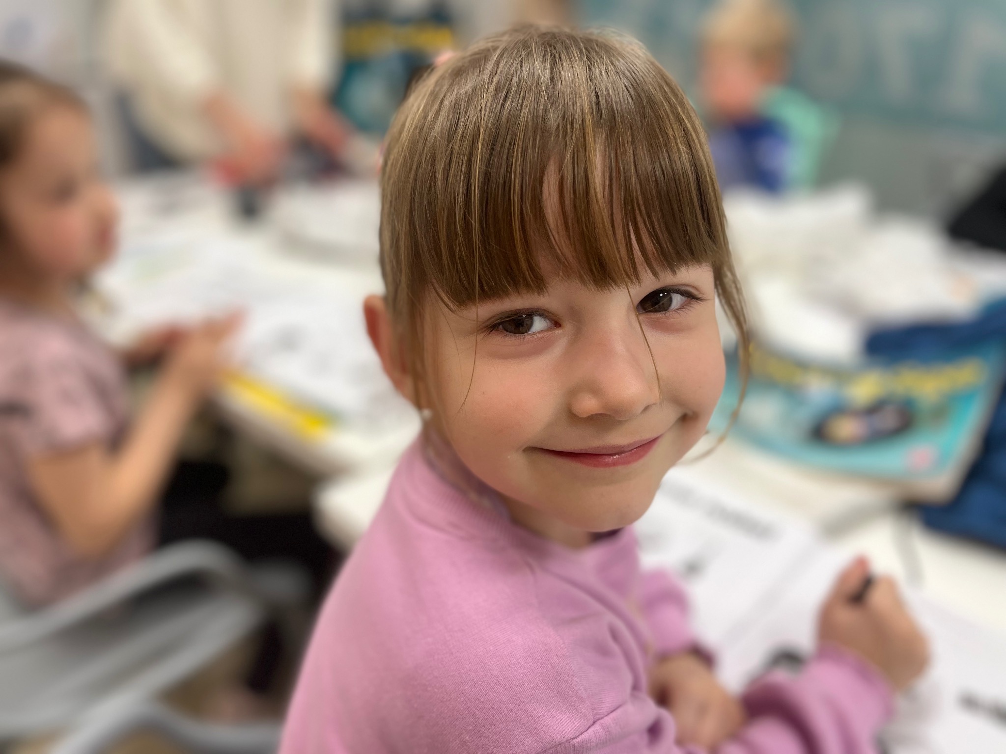 Uśmiechnięta dziewczynka w różowej bluzce siedzi przy stole z innymi dziećmi, prawdopodobnie podczas lekcji języka obcego. W dłoni trzyma długopis.