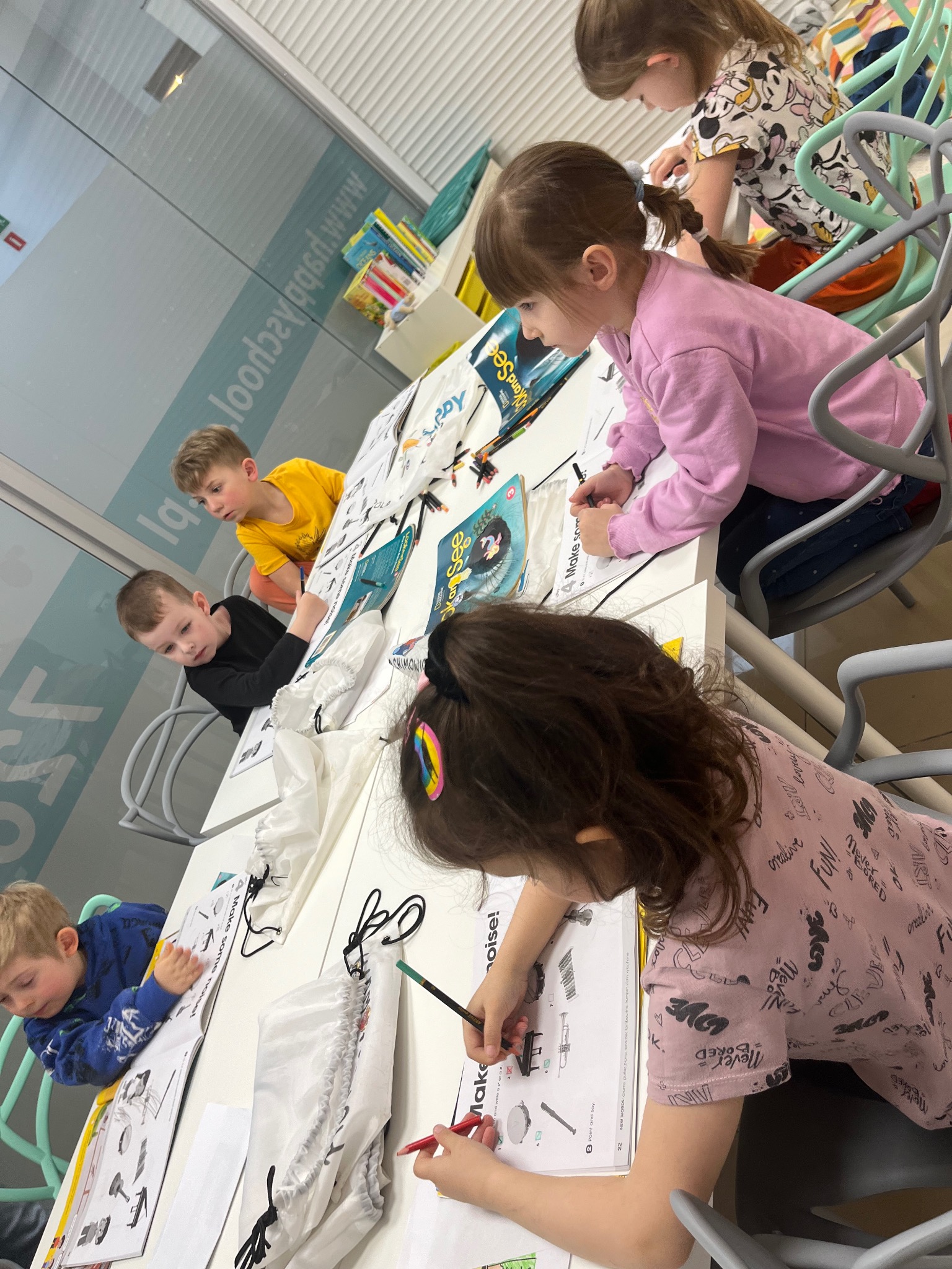 Dzieci w jasnej sali uczą się słówek, rysując instrumenty muzyczne z książki 'Make some noise!'. Na stole leżą torby na buty. Lekcja w szkole językowej.