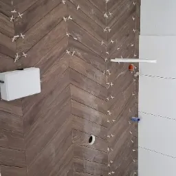 Montaż płytek imitujących drewno w jodełkę na ścianie w łazience, z widocznymi krzyżykami dystansowymi i instalacją hydrauliczną w trakcie prac.