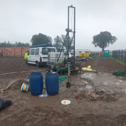 Wiercenie studni w błotnistym terenie; widoczna wiertnica, biały van z rurami na dachu, agregat prądotwórczy na przyczepce oraz dwa niebieskie beczki.