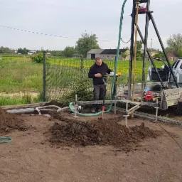 Wiercenie studni głębinowej na działce, widoczny pracownik obsługujący wiertnicę, wykopany urobek oraz samochód dostawczy z przyczepą.