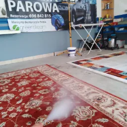 Czyszczenie parowe czerwonego dywanu z orientalnym wzorem za pomocą niebieskiego urządzenia parowego; w tle baner reklamowy myjni parowej i regał z akcesoriami.