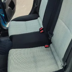 Wnętrze błękitnego samochodu, widoczna tylna kanapa z jasną, wzorzystą tapicerką i czarnymi bokami, pasy bezpieczeństwa z czerwonymi zatrzaskami oraz gumowe dywaniki podłogowe.