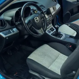 Wnętrze błękitnej Mazdy 3 z jasną tapicerką siedzeń i czarną deską rozdzielczą, widoczny odświeżacz powietrza Wunder-Baum zwisający z lusterka.